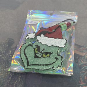May include: A textured light green character ornament with a dark red Santa hat and white trim, featuring yellow eyes and black outlines. The ornament is enclosed in a clear, iridescent plastic bag with a black hanging cord and small beads.
