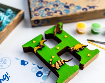 Könnte beinhalten: Ein grüner Holzbuchstabe H, der mit kleinen Dinosauriern und Fledermäusen aus Holz verziert ist. Der Buchstabe ist von Bastelutensilien umgeben, darunter Farbe, ein kleiner Schraubendreher und goldener Glitzer.