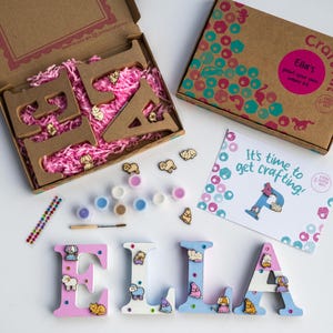May include: A craft kit for children to decorate wooden letters with paint and embellishments. The kit includes the letters "ELLA", paint, embellishments, and a craft card that says "It's time to get crafting!"