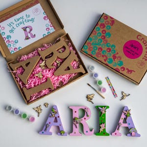 May include: A craft kit for decorating wooden letters with fairy and flower designs. The kit includes paints, glitter, and embellishments. The letters spell out the name "ARIA".