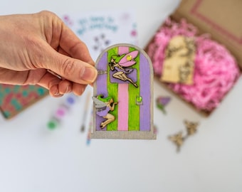 Puede incluir: Una puerta de hadas de madera con un diseño a rayas de color morado y verde. La puerta presenta dos hadas con alas, una volando y otra de pie. La puerta está pintada con un patrón de rayas de color rosa claro y verde.