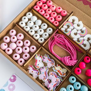 May include: A wooden box filled with colourful beads, wooden unicorn charms, rainbow charms, and elastic string. The box is labelled "Little Crafty Bees" and "Show us your art!"