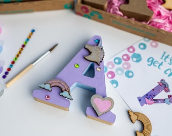 Puede incluir: Letra A de madera de color morado decorada con un arcoíris, un unicornio y un corazón. La letra forma parte de un kit de manualidades que incluye pintura, pedrería y una tarjeta que dice "Es hora de hacer manualidades".