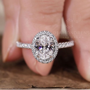 Może przedstawiać: Srebrny pierścionek zaręczynowy z dużym, owalnym diamentem otoczonym halo z mniejszych diamentów. Pierścionek ma cienki obrączek z małymi diamentami osadzonymi po bokach.