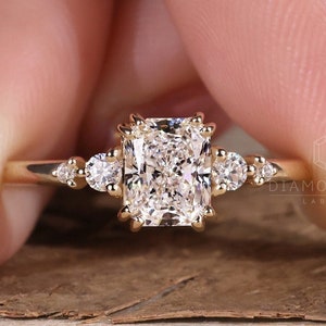 Könnte beinhalten: Ein goldener Verlobungsring mit einem großen, quadratischen Diamanten als Mittelstück und kleineren Akzentdiamanten auf der Ringschiene. Der Ring liegt auf einer Holzoberfläche.