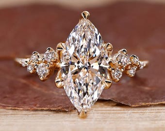 Op de afbeelding: Een gouden verlovingsring met een marquise-geslepen diamant. De ring heeft een grote centrale diamant en kleinere diamanten aan weerszijden. De ring ligt op een bruin blad.