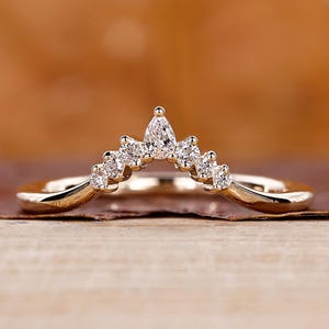 Könnte beinhalten: Ein zarter Goldring mit geschwungenem Band, besetzt mit kleinen, funkelnden Diamanten. Die Diamanten sind in einem anmutigen, nach oben gerichteten Bogen angeordnet und bilden ein kronenartiges Design. Der Ring wird auf einer Holzoberfläche präsentiert.