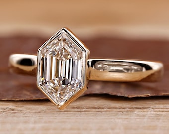 May include: A gold ring with a unique, geometric-cut diamond. The diamond is a clear, multifaceted stone set in a gold band. The ring is displayed on a wooden surface, showcasing its elegant design.