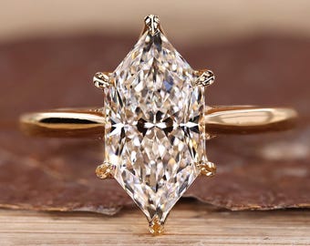 May include: A gold engagement ring featuring a large, clear, marquise-cut diamond. The diamond is set with four prongs and sits atop a thin, polished gold band. The ring is displayed on a wooden surface.