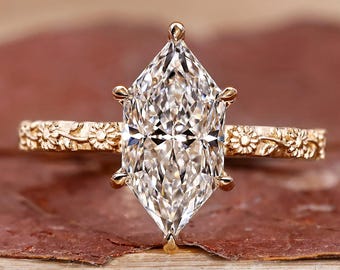 May include: A gold engagement ring with a large, clear, marquise-cut diamond. The band features a floral design. The ring is set on a reddish-brown surface.
