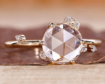 May include: A gold ring with a large, clear, faceted gemstone as the centerpiece. Small accent stones are clustered around the main stone and along the band, creating a delicate, nature-inspired design. The ring is set against a warm, neutral background.