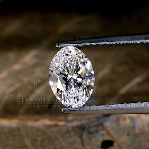 Może przedstawiać: Przezroczysty diament o owalnym kształcie trzymany w pincecie na tle brązowego liścia. Diament jest hodowany w laboratorium i ma szlif brylantowy.