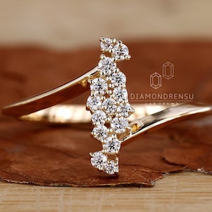 May include: A gold ring with a unique design featuring a cluster of small diamonds set in a curved shape. The ring is on a wooden surface. DIAMONDRENSU LAB GROWN is printed on the ring.