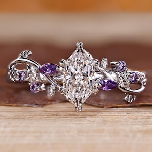 May include: A silver ring featuring a large, marquise-cut clear gemstone at the center, flanked by smaller purple gemstones and delicate floral accents. The ring is set against a warm, wooden background, highlighting its intricate design.