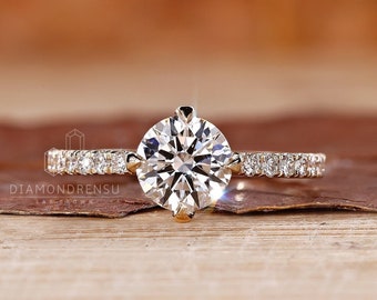 May include: A gold engagement ring with a round brilliant cut diamond center stone and a pave diamond band. The ring is set on a wooden surface.