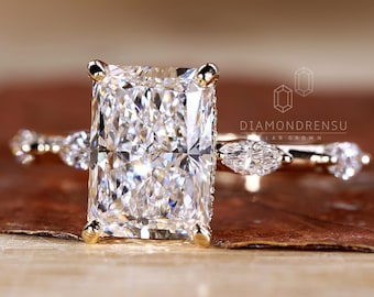 Puede incluir: Un anillo de compromiso de oro con un gran diamante central rectangular y transparente. La banda está adornada con diamantes más pequeños en forma de pera a cada lado del diamante central. El anillo está colocado sobre una superficie marrón texturizada. DIAMONDRENSU LAB GROWN.