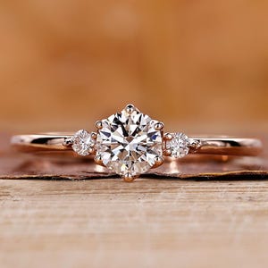 Könnte beinhalten: Ein Verlobungsring aus Roségold mit einem großen runden Diamanten in der Mitte und zwei kleineren Akzentdiamanten auf jeder Seite.