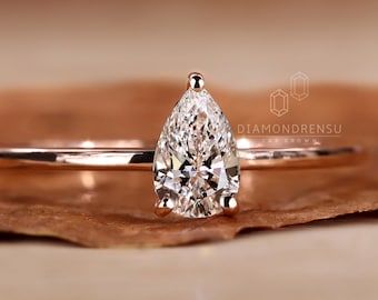 May include: A pear-shaped diamond engagement ring set in a rose gold band. The ring is on a brown background.