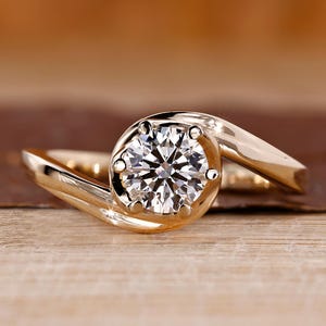 May include: A gold engagement ring with a large, round, clear diamond center stone. The band curves around the diamond in a swirling design. The ring is set against a blurred wooden background.