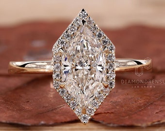 Può includere: Un anello di fidanzamento in oro con una grande pietra centrale di diamante taglio marquise, circondata da un alone di diamanti più piccoli. L'anello ha una fascia delicata e un design ad alone geometrico unico. L'anello è su una superficie marrone.