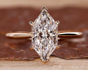 May include: A gold engagement ring with a marquise-cut diamond. The diamond is set with four prongs and has a brilliant cut. The ring band is thin and polished. The ring is set on a wooden surface.