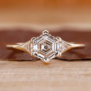 May include: A gold ring featuring a large, hexagonal-cut clear gemstone as the centerpiece. The ring has a delicate band and smaller triangular accent stones on either side of the main stone. The ring is set against a warm, wooden background.