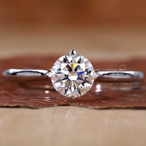 K&ouml;nnte beinhalten: Ein Solit&auml;r-Verlobungsring mit einem gro&szlig;en, runden Diamanten im Brillantschliff, der in einem schlanken, silbernen Band gefasst ist. Der Diamant funkelt hell. Der Ring wird auf einer braunen Oberfl&auml;che pr&auml;sentiert.