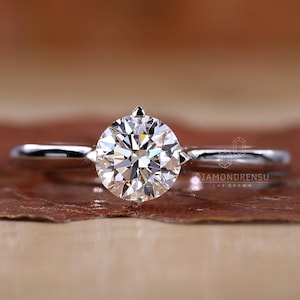 K&ouml;nnte beinhalten: Ein Solit&auml;r-Verlobungsring mit einem runden, brillantgeschliffenen Diamanten, der in einem polierten Silberband gefasst ist. Der Diamant ist der Mittelpunkt und reflektiert das Licht. Der Ring wird auf einer braunen Oberfl&auml;che pr&auml;sentiert.