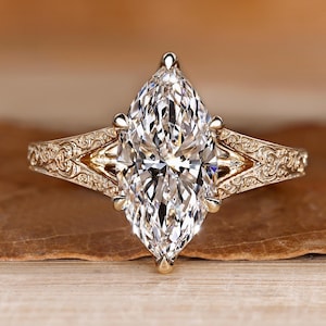 Puede incluir: Un anillo con una piedra preciosa transparente de corte marquesa montado en una banda de oro amarillo. La banda presenta intrincados patrones de volutas grabados a lo largo de los hombros, que conducen a un engaste de seis garras.