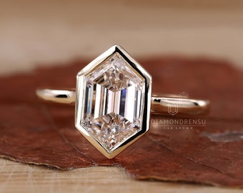 May include: A gold engagement ring with a hexagonal bezel setting featuring a clear, octagonal-shaped diamond. The ring is set on a brown wooden surface. "DIAMONDRENSU LAB GROWN" is printed on the surface.