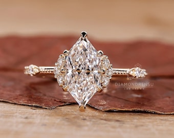 Puede incluir: Un anillo de compromiso de oro con un gran diamante central de forma marquesa, claro y brillante. La piedra central está rodeada de diamantes más pequeños engastados en un halo. La banda es delicada y presenta un diseño de grano de mijo.
