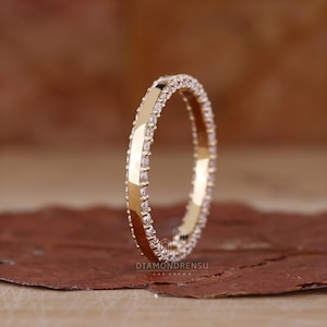 Könnte beinhalten: Ein dünner Goldring mit einer Reihe kleiner Diamanten, die um den Rand herumgesetzt sind. Der Ring ist glatt und poliert, und die Diamanten sind klar und funkelnd.