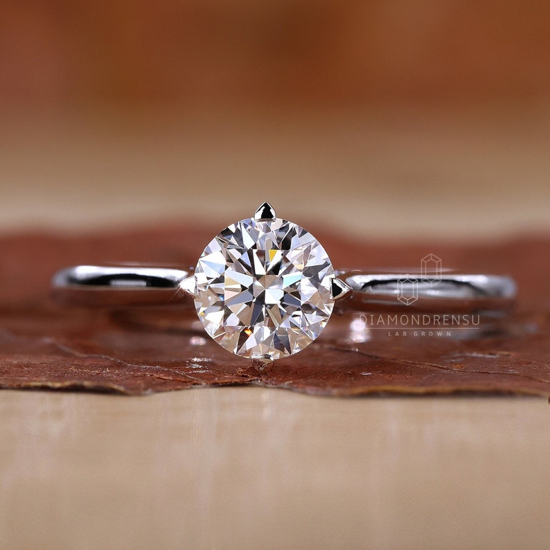 K&ouml;nnte beinhalten: Ein Solit&auml;r-Verlobungsring mit einem runden, brillantgeschliffenen Diamanten, der in einem polierten Silberband gefasst ist. Der Diamant ist der Mittelpunkt und reflektiert das Licht. Der Ring wird auf einer braunen Oberfl&auml;che pr&auml;sentiert.