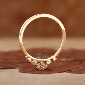 K&ouml;nnte beinhalten: Ein zarter Goldring mit einem kleinen, runden Diamanten in der Mitte und kleineren Diamanten, die entlang der Ringschiene gesetzt sind. Der Ring liegt auf einer braunen Oberfl&auml;che.