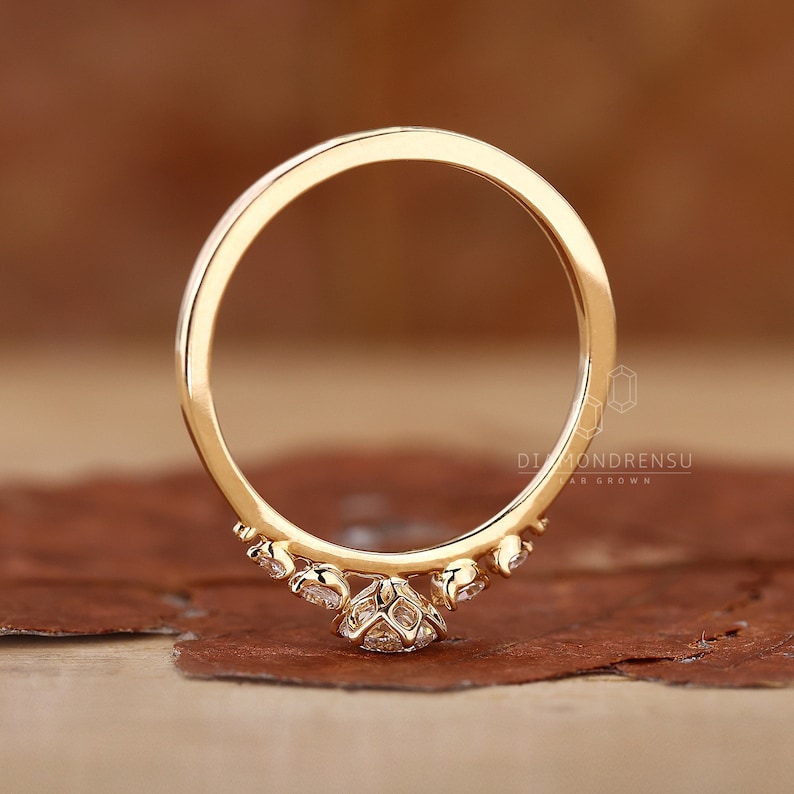 K&ouml;nnte beinhalten: Ein zarter Goldring mit einem kleinen, runden Diamanten in der Mitte und kleineren Diamanten, die entlang der Ringschiene gesetzt sind. Der Ring liegt auf einer braunen Oberfl&auml;che.
