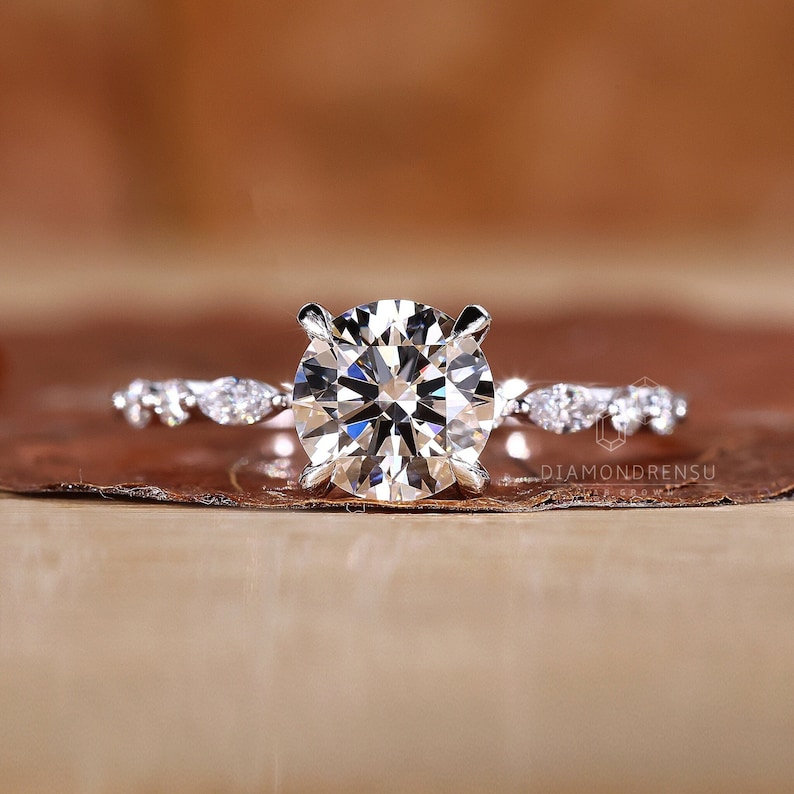 May include: A silver engagement ring with a large round diamond center stone and a delicate band of smaller diamonds. The ring is set on a brown background.