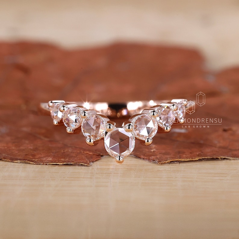 May include: Rose gold wedding band with a curved row of round, bezel-set, rose cut diamonds. The band is made of 14k rose gold and features a total of 9 diamonds.