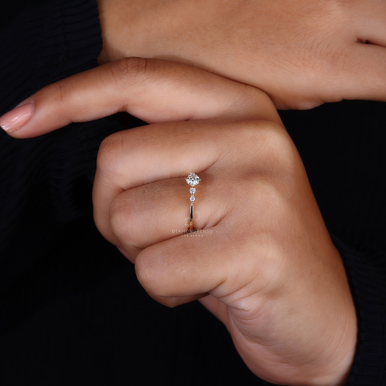 K&ouml;nnte beinhalten: Ein zarter Goldring mit einem runden Diamanten in der Mitte und kleineren Diamanten auf der Ringschiene. Der Ring befindet sich an einem Finger.