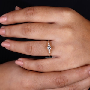 K&ouml;nnte beinhalten: Ein zarter Goldring mit einem runden Diamanten in der Mitte und kleineren Diamanten auf der Ringschiene. Der Ring befindet sich an einem Finger.