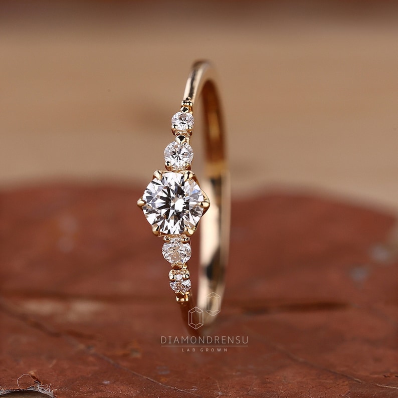 K&ouml;nnte beinhalten: Ein zarter Goldring mit einem runden Diamanten in der Mitte und kleineren Diamanten, die entlang der Ringschiene gesetzt sind. Der Ring liegt auf einem braunen Hintergrund.