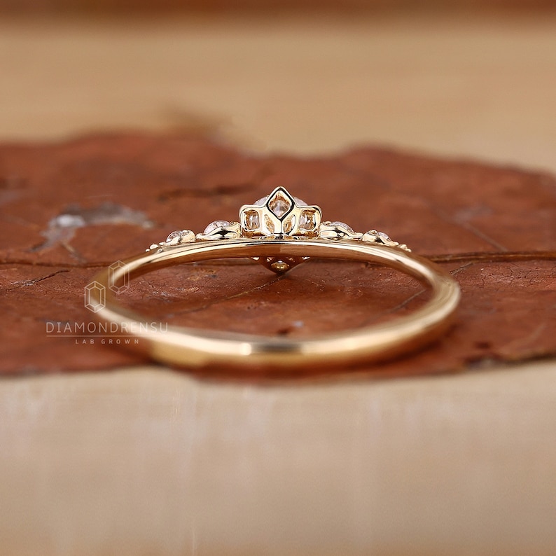 K&ouml;nnte beinhalten: Ein zarter Goldring mit einem kleinen, klaren Diamanten in der Mitte und kleineren Diamanten auf der Ringschiene. Der Ring liegt auf einer braunen, strukturierten Oberfl&auml;che.