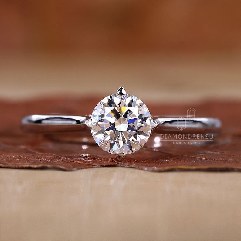 K&ouml;nnte beinhalten: Ein Solit&auml;r-Verlobungsring mit einem gro&szlig;en, runden Diamanten im Brillantschliff, der in einem schlanken, silbernen Band gefasst ist. Der Diamant funkelt hell. Der Ring wird auf einer braunen Oberfl&auml;che pr&auml;sentiert.