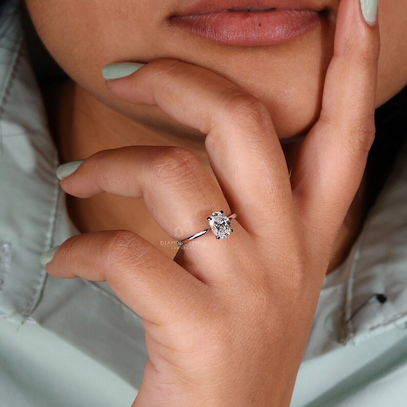 May include: A silver solitaire engagement ring with an oval-shaped diamond. The ring is on a person's finger.