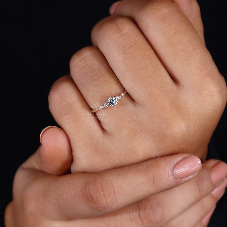 K&ouml;nnte beinhalten: Ein zarter Goldring mit einem Cluster aus kleinen Diamanten, die in einem geschwungenen Band gefasst sind. Der Ring befindet sich an einer Hand mit hellrosa Nagellack.