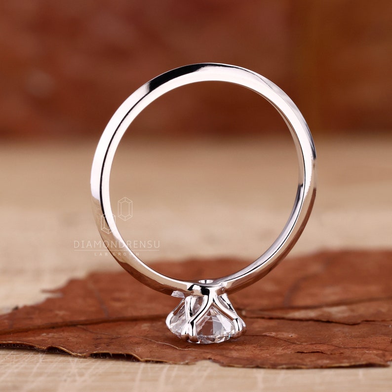Puede incluir: Un anillo de compromiso solitario de plata con un diamante redondo brillante engastado en una garra de cuatro puntas.