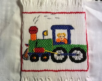 Puede incluir: Un cuadrado de tela blanca con un diseño de tren bordado de colores. La tela tiene un borde rizado. La imagen también incluye una etiqueta blanca con el texto "Item #:" "080" "Description:" "train engine" y "QTY:".