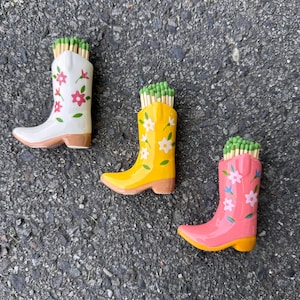 May include: Three ceramic cowboy boot match holders in white, yellow, and pink, each filled with green-tipped matches. The boots are decorated with floral designs and have brown heels.