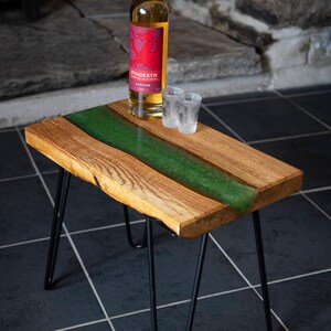 May include: A wooden table with a green epoxy resin river running down the center. The table has black hairpin legs and is sitting on a black tiled floor. A bottle of Penderyn Legend Welsh whisky and two shot glasses are on the table.