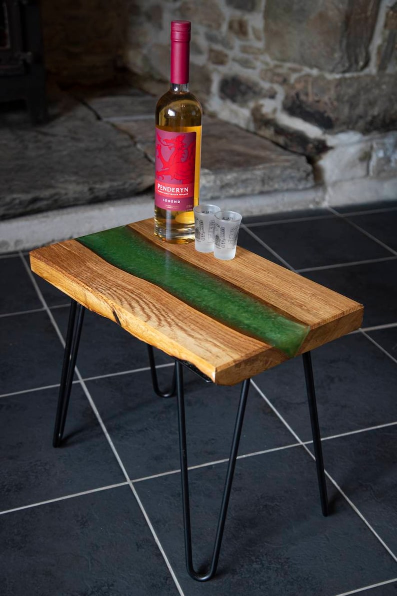 May include: A wooden table with a green epoxy resin river running down the center. The table has black hairpin legs and is sitting on a black tiled floor. A bottle of Penderyn Legend Welsh whisky and two shot glasses are on the table.
