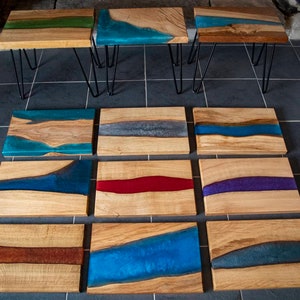 May include: A collection of 10 wooden serving trays with various colored resin river designs. The trays are arranged in three rows, with the top row featuring three trays on black hairpin legs. The resin designs include blue, green, gray, red, purple, and brown.